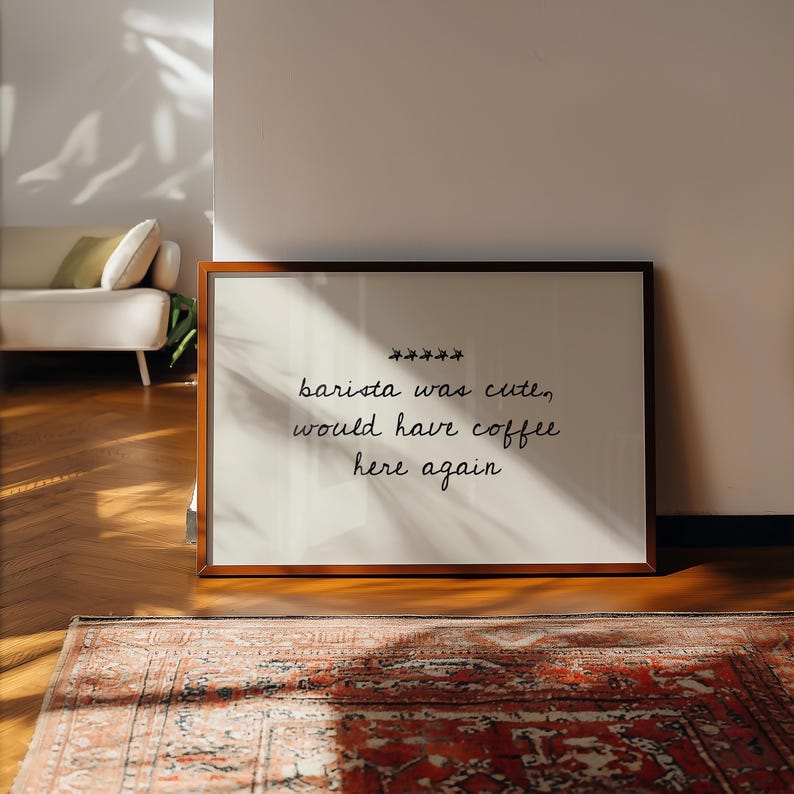 May include: A framed print with the text "barista was cute, would have coffee here again" in a handwritten style. The print is in a brown wooden frame, resting on a wooden floor with a patterned rug in the foreground.