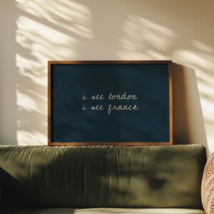 May include: A framed art print with the text "i see london i see france" in a handwritten style. The artwork has a dark blue background and a wooden frame. The print is displayed on a green sofa.