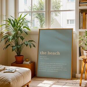 Puede incluir: Un p&oacute;ster enmarcado con el texto "the beach" en blanco sobre un fondo azul claro. El p&oacute;ster est&aacute; apoyado contra una pared en una habitaci&oacute;n con plantas, una mesa auxiliar de madera y una estanter&iacute;a. Una planta grande en una maceta de terracota est&aacute; en primer plano.