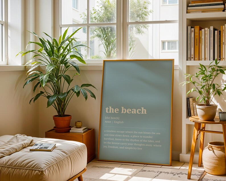 Puede incluir: Un p&oacute;ster enmarcado con el texto "the beach" en blanco sobre un fondo azul claro. El p&oacute;ster est&aacute; apoyado contra una pared en una habitaci&oacute;n con plantas, una mesa auxiliar de madera y una estanter&iacute;a. Una planta grande en una maceta de terracota est&aacute; en primer plano.
