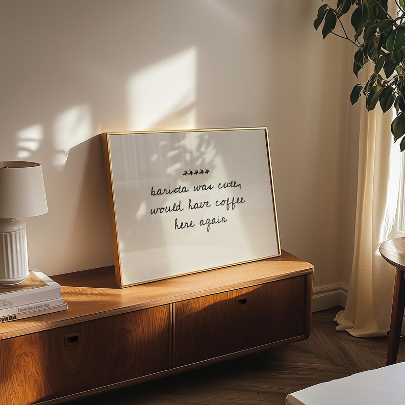 May include: A framed print with the text "barista was cute, would have coffee here again" in black script. The print has a gold frame and is displayed on a wooden cabinet. A lamp and books are visible on the cabinet.