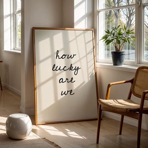 May include: A framed print with the words "how lucky are we" in a light-filled room. A wooden chair, a potted plant, and a small white stool are also visible. The artwork is leaning against a wall.
