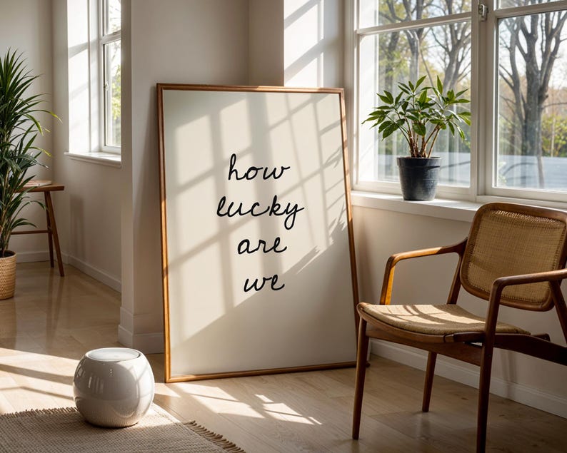 May include: A framed print with the words "how lucky are we" in a light-filled room. A wooden chair, a potted plant, and a small white stool are also visible. The artwork is leaning against a wall.
