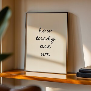 May include: A framed art print with the words "how lucky are we" in black script. The print has a cream-coloured background and a thin gold frame. It rests on a wooden shelf with a stack of black books, near a window with a green plant.