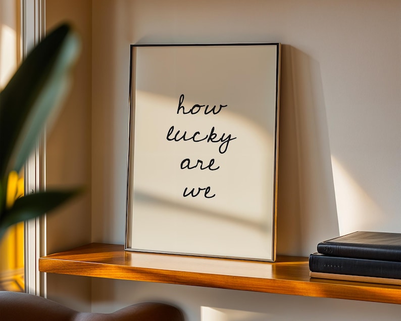 May include: A framed art print with the words "how lucky are we" in black script. The print has a cream-coloured background and a thin gold frame. It rests on a wooden shelf with a stack of black books, near a window with a green plant.