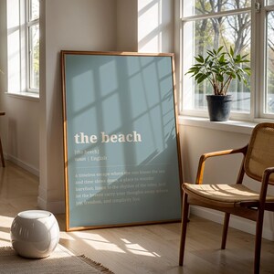 Puede incluir: Un p&oacute;ster enmarcado con las palabras "the beach" en una fuente clara sobre un fondo azul p&aacute;lido. La habitaci&oacute;n incluye una silla de madera, una peque&ntilde;a planta en maceta y un taburete de cer&aacute;mica blanco. La habitaci&oacute;n tiene grandes ventanas.