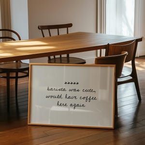May include: A framed white sign with the text "barista was cute, would have coffee here again" in black script. The sign is in a light wood frame and is placed in front of a wooden table and chairs.