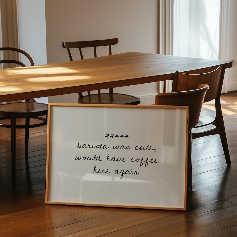 May include: A framed white sign with the text "barista was cute, would have coffee here again" in black script. The sign is in a light wood frame and is placed in front of a wooden table and chairs.