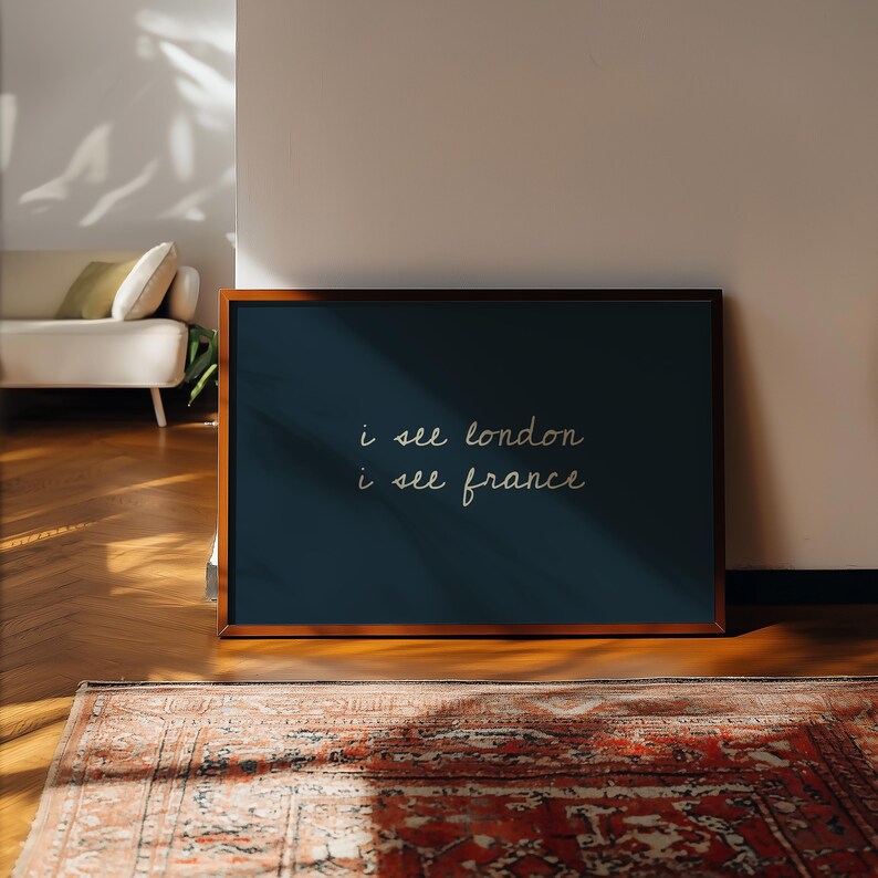 May include: A framed artwork with the text "i see london i see france" in white script on a dark blue background. The frame is a warm brown. The artwork is on a wooden floor, with a patterned rug in the foreground.