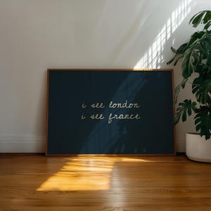 May include: A framed art print with the text "i see london i see france" in a handwritten style. The artwork has a dark teal background and a wooden frame. The print is displayed on a wooden floor with sunlight.