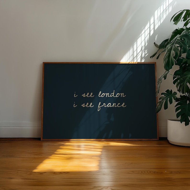 May include: A framed art print with the text "i see london i see france" in a handwritten style. The artwork has a dark teal background and a wooden frame. The print is displayed on a wooden floor with sunlight.