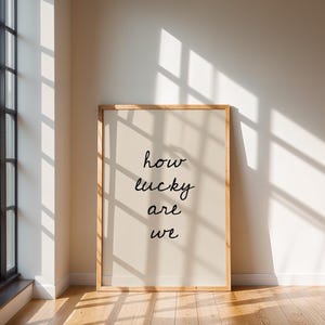 May include: A framed print with the words "how lucky are we" in black script. The print has a light beige background and a natural wood frame. The image is set against a white wall with sunlight streaming through a window.