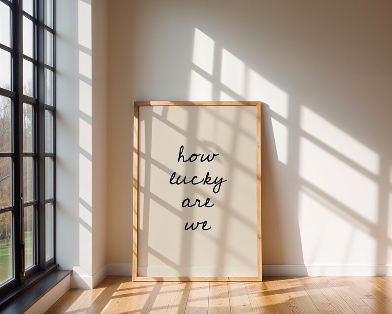 May include: A framed print with the words "how lucky are we" in black script. The print has a light beige background and a natural wood frame. The image is set against a white wall with sunlight streaming through a window.