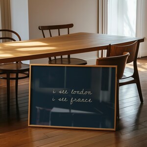 May include: A framed navy blue print with the text "i see london i see france" in a cursive font. The print is displayed on a wooden floor, with a wooden table and chairs in the background. The frame is a light brown colour.
