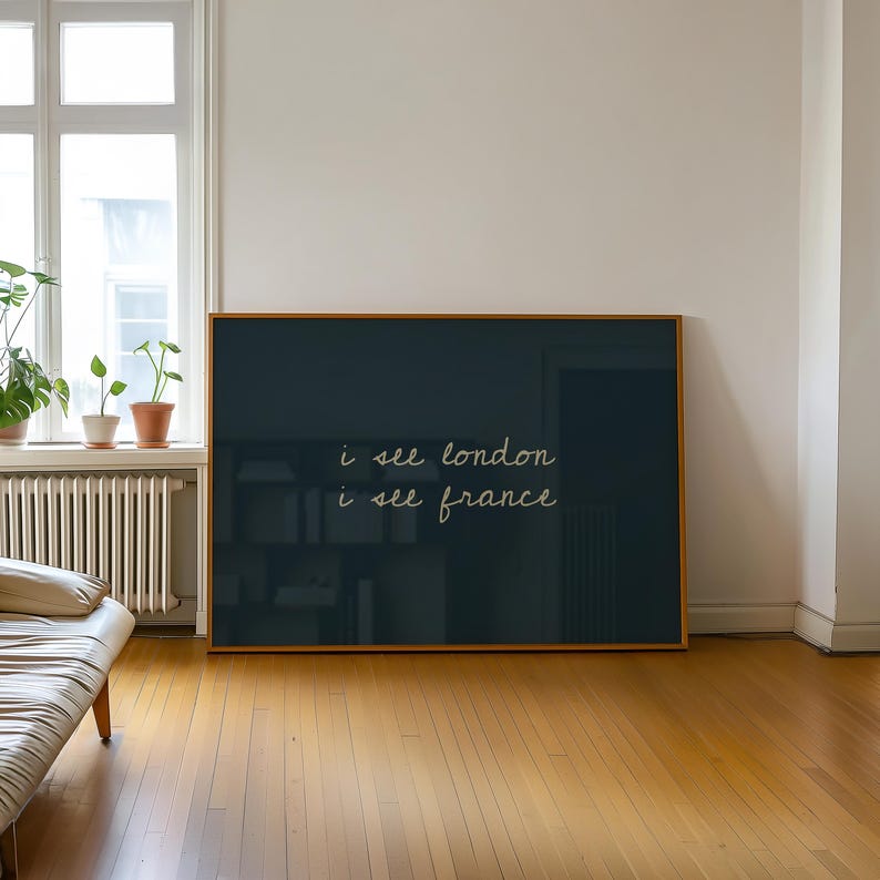 May include: A framed art print with a dark teal background and the handwritten text "i see london i see france" in a cursive font. The frame is a warm, light brown colour, and the artwork is displayed in a room with hardwood floors.