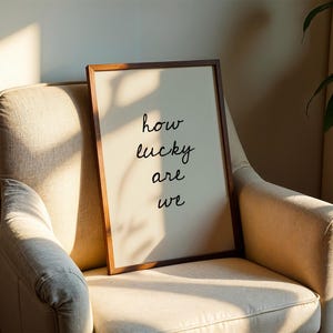 May include: A framed print with the words "how lucky are we" in black script. The print is in a brown wooden frame and leans against a beige armchair. The scene is lit by natural light, creating shadows.