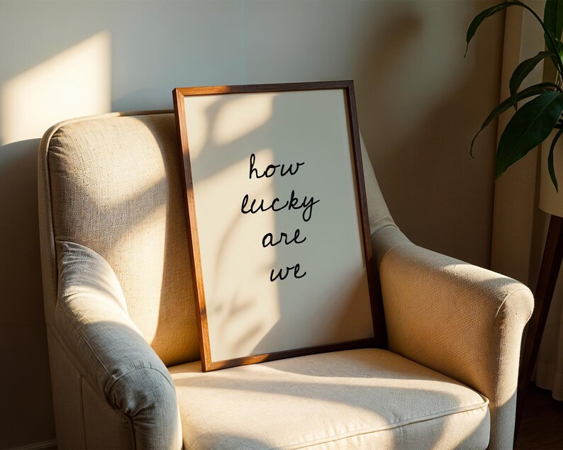 May include: A framed print with the words "how lucky are we" in black script. The print is in a brown wooden frame and leans against a beige armchair. The scene is lit by natural light, creating shadows.