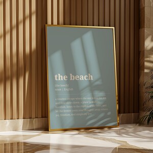 Puede incluir: Un marco dorado contra una pared de madera. El cartel muestra la palabra "the beach" en blanco sobre un fondo azul claro. El texto incluye una definici&oacute;n de la palabra, en ingl&eacute;s.
