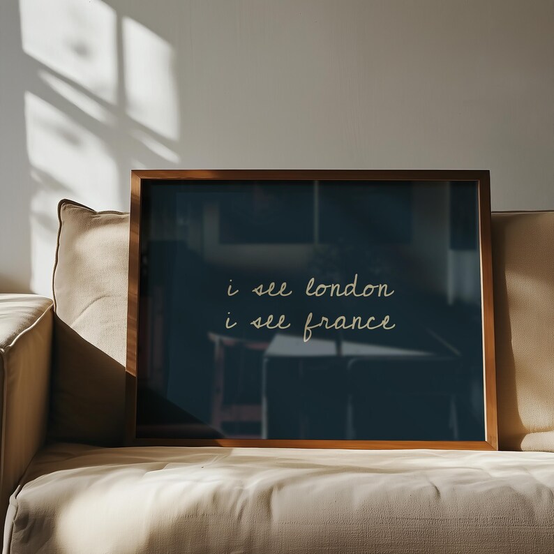 May include: A framed print featuring the handwritten text "i see london i see france". The frame is dark wood, and the background is a blurred interior scene. The print is displayed on a beige sofa, with sunlight casting shadows.