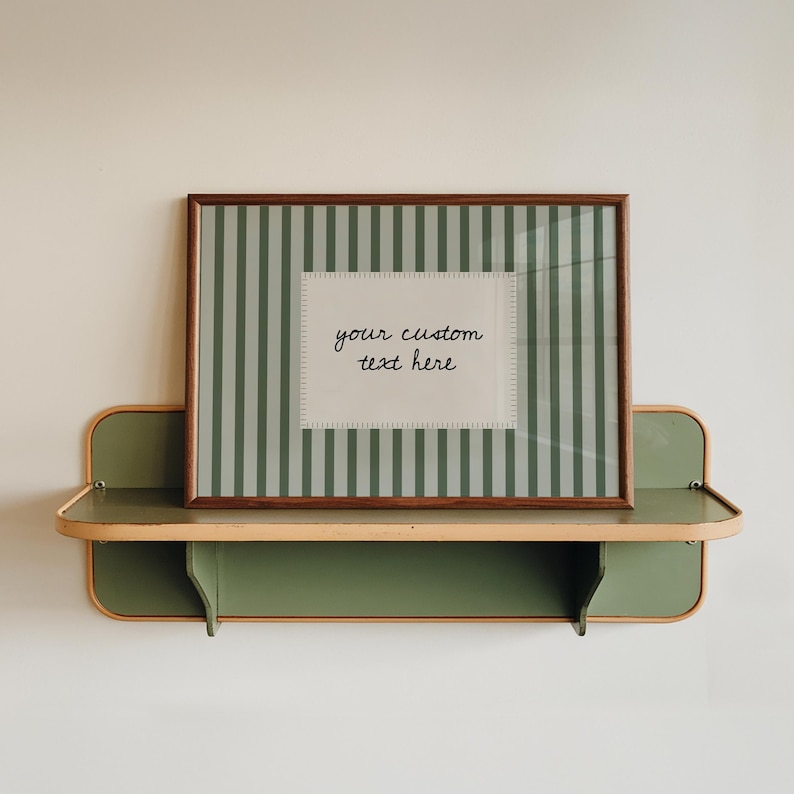 Pu&ograve; includere: Una cornice rettangolare con finitura in legno scuro contenente una stampa a righe verticali verde salvia e bianche, con un'etichetta rettangolare bianca centrale che riporta il testo scritto a mano in nero your custom text here.
