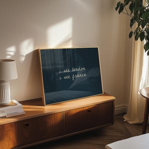 May include: A framed art print with a gold frame, featuring the text "i see london i see france" in white script on a dark blue background. The print is displayed on a wooden cabinet in a well-lit room.