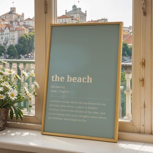 Puede incluir: Una impresi&oacute;n enmarcada con las palabras "the beach" en texto blanco sobre un fondo azul claro. La impresi&oacute;n est&aacute; en un marco dorado y est&aacute; apoyada contra un alf&eacute;izar con vistas a una ciudad.