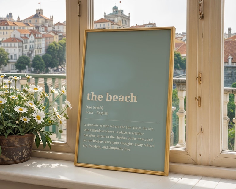 Puede incluir: Una impresi&oacute;n enmarcada con las palabras "the beach" en texto blanco sobre un fondo azul claro. La impresi&oacute;n est&aacute; en un marco dorado y est&aacute; apoyada contra un alf&eacute;izar con vistas a una ciudad.