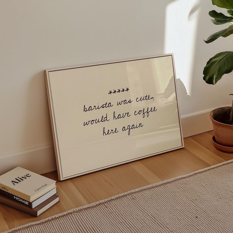 May include: A framed print with the text "barista was cute, would have coffee here again" in black script. The frame is a light brown colour. Several books are stacked on the floor in front of the print.