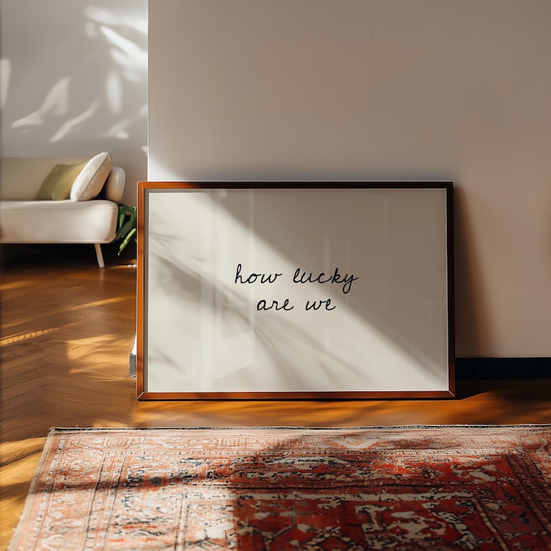 May include: A framed print with the words "how lucky are we" in a handwritten style. The frame is a dark wood color, and the print is leaning against a wall. A rug with a red and brown pattern is in the foreground.