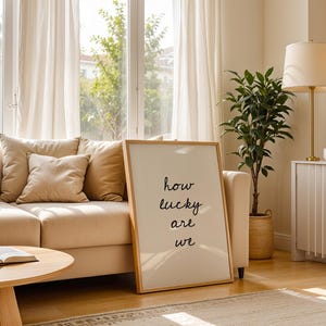 May include: A bright living room featuring a beige sofa with cushions, a wooden coffee table, and a framed print with the words "how lucky are we". A floor lamp, a potted plant, and a window with curtains create a warm, inviting atmosphere.
