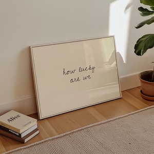 May include: A framed print with the text "how lucky are we" in black script. The print has a cream-colored background and a thin, gold-colored frame. The print is leaning against a white wall, next to a stack of books.
