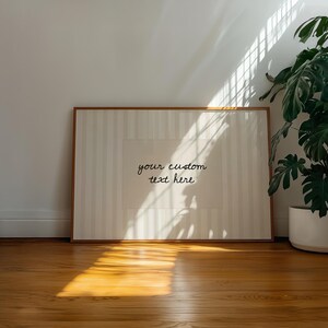 Pu&ograve; includere: Una stampa rettangolare incorniciata con uno sfondo beige chiaro a righe verticali e un'area centrale rettangolare contenente il testo corsivo nero your custom text here. La cornice &egrave; realizzata in legno chiaro sottile.