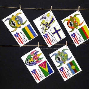 May include: Five colorful cards with a stylized monster face and a flag on each card. The cards are hanging on a string with clothespins. The text on the cards reads "Hatillo, Puerto Rico", "Bayamón, Puerto Rico", "Humosae, Puerto Rico", "Moca, Puerto Rico", and "Utuado, Puerto Rico".