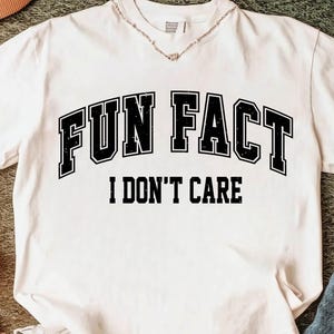 May include: White t-shirt with the text "FUN FACT I DON'T CARE" in black block letters. The shirt has a V-neck and a casual style, suitable for everyday wear. A necklace is visible at the neckline.