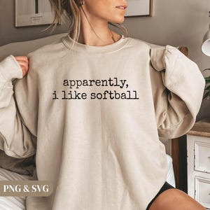 K&ouml;nnte beinhalten: Beiges Sweatshirt mit dem schwarzen Text "apparently, i like softball". Das Sweatshirt hat einen Rundhalsausschnitt und lange &Auml;rmel. Das Bild zeigt eine Nahaufnahme des Sweatshirts.