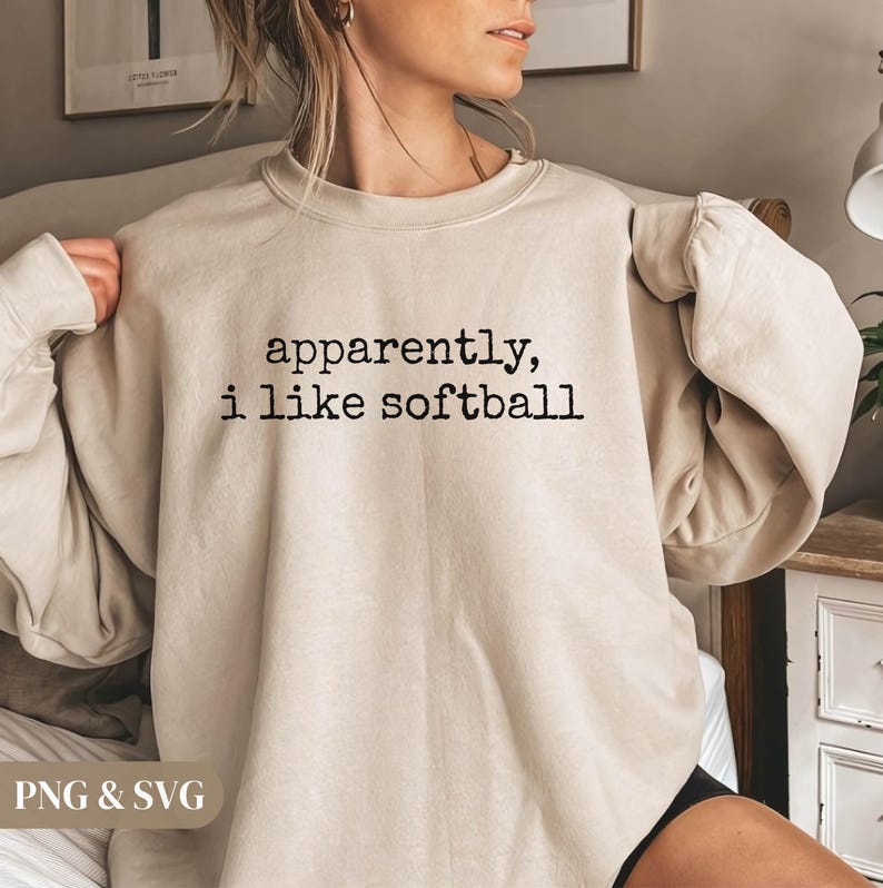 K&ouml;nnte beinhalten: Beiges Sweatshirt mit dem schwarzen Text "apparently, i like softball". Das Sweatshirt hat einen Rundhalsausschnitt und lange &Auml;rmel. Das Bild zeigt eine Nahaufnahme des Sweatshirts.