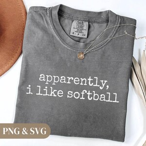 K&ouml;nnte beinhalten: Ein graues Comfort Colors T-Shirt mit dem wei&szlig;en Text "apparently, i like softball". Eine silberne Halskette mit einem kleinen Anh&auml;nger liegt auf dem Shirt. Ein brauner Hut ist oben links zu sehen.