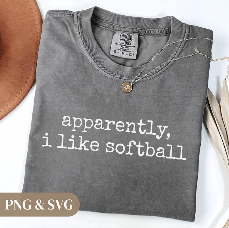 K&ouml;nnte beinhalten: Ein graues Comfort Colors T-Shirt mit dem wei&szlig;en Text "apparently, i like softball". Eine silberne Halskette mit einem kleinen Anh&auml;nger liegt auf dem Shirt. Ein brauner Hut ist oben links zu sehen.
