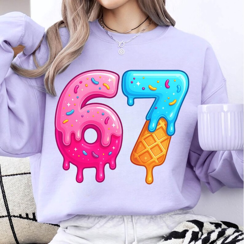 May include: A light purple sweatshirt featuring a colorful graphic of the number "67". The "6" is designed as a pink donut with sprinkles, while the "7" is blue and dripping, resting on a waffle cone.