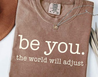 May include: A brown t-shirt with the words "be you. the world will adjust" printed in white. A silver necklace with a small pendant rests on the shirt. A brown hat is visible in the upper left corner.