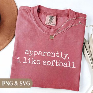 K&ouml;nnte beinhalten: Ein staubrosafarbenes T-Shirt mit dem wei&szlig;en Aufdruck "apparently, i like softball". Eine silberne Halskette mit einem kleinen Anh&auml;nger liegt auf dem Shirt. Das Shirt ist mit "Comfort Colors" gekennzeichnet und hat die Gr&ouml;&szlig;e S.