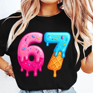 May include: Black t-shirt featuring the number "67" in a playful, colorful design. The "6" is pink with sprinkles and a donut shape, while the "7" is blue with a waffle cone base. A fun, celebratory graphic.