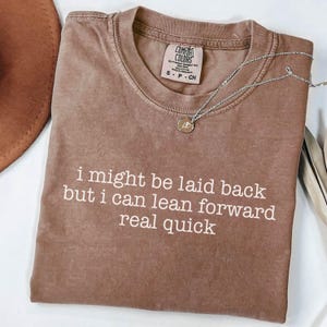 May include: A brown Comfort Colors t-shirt with the text "i might be laid back but i can lean forward real quick" in white. A silver necklace with a small pendant is on the shirt.
