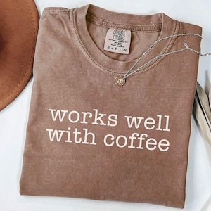 May include: A brown Comfort Colors t-shirt with the phrase "works well with coffee" in white. A silver necklace with a small pendant rests on the shirt. A brown hat is partially visible in the upper left corner.