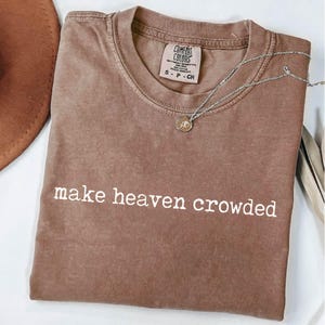 May include: A brown t-shirt with the phrase "make heaven crowded" printed in white. A silver necklace with a small pendant is draped across the shirt. The shirt has a tag that reads "Comfort Colors".