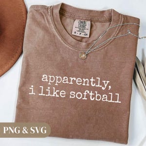 K&ouml;nnte beinhalten: Ein braunes T-Shirt mit dem wei&szlig;en Text "apparently, i like softball". Das Shirt hat einen Rundhalsausschnitt und ein kleines Etikett oben. Eine silberne Halskette mit Anh&auml;nger liegt auf dem Shirt.