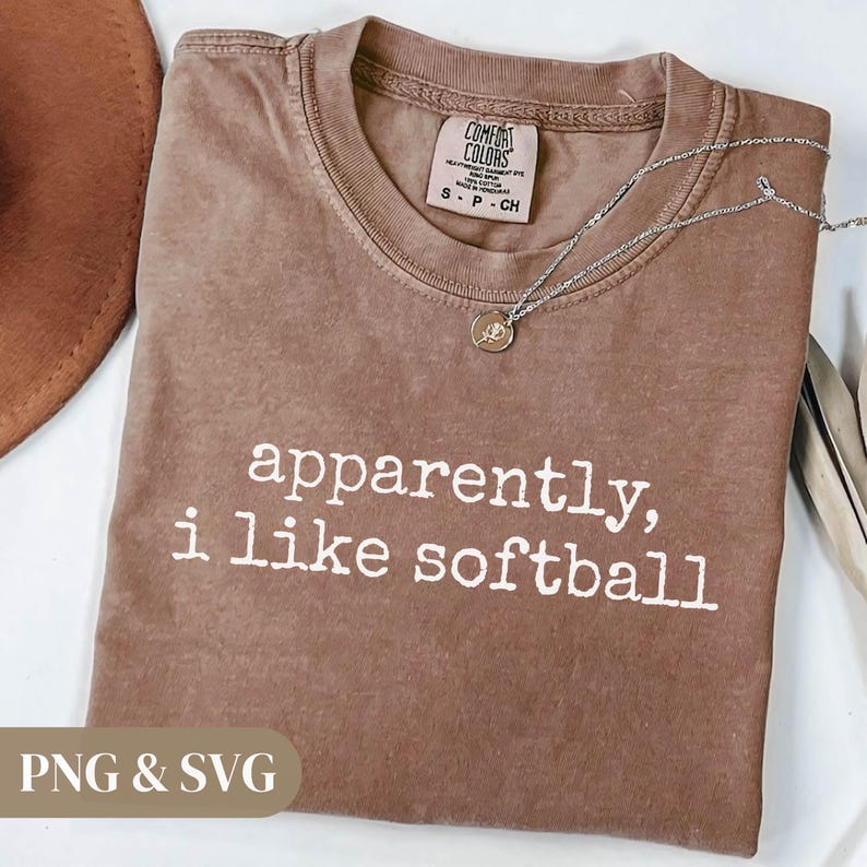 K&ouml;nnte beinhalten: Ein braunes T-Shirt mit dem wei&szlig;en Text "apparently, i like softball". Das Shirt hat einen Rundhalsausschnitt und ein kleines Etikett oben. Eine silberne Halskette mit Anh&auml;nger liegt auf dem Shirt.
