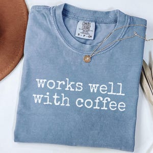 May include: A blue Comfort Colors t-shirt with the phrase "works well with coffee" in white lettering. A gold necklace with a small pendant rests on the shirt. A brown hat is visible in the upper left corner.