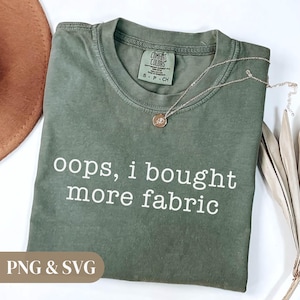 Puede incluir: Una camiseta verde oliva con el texto "oops, i bought more fabric" en blanco. La camiseta tiene cuello redondo y mangas cortas. Un collar dorado con un colgante está drapeado sobre la camiseta. Un sombrero marrón y plantas secas en el fondo.