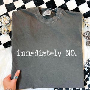 Puede incluir: Una sudadera gris de cuello redondo con las palabras "immediately NO." impresas en blanco. La camisa está doblada y se muestra sobre una superficie a cuadros en blanco y negro. La camisa tiene un aspecto vintage y desgastado.